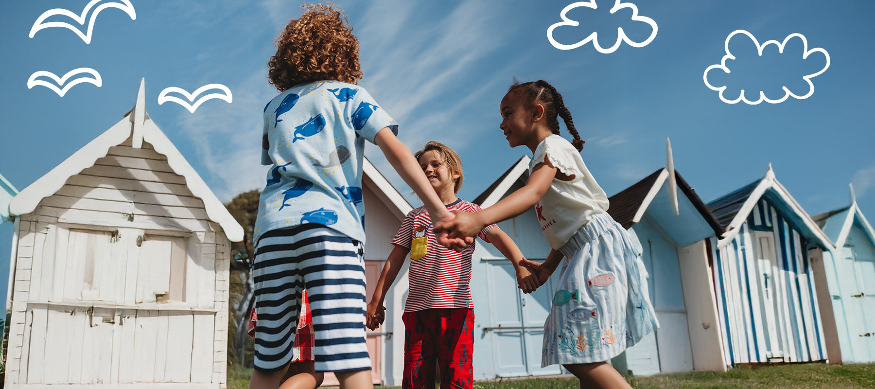Children holding hands in front of beach huts with a blue sky and clouds.
